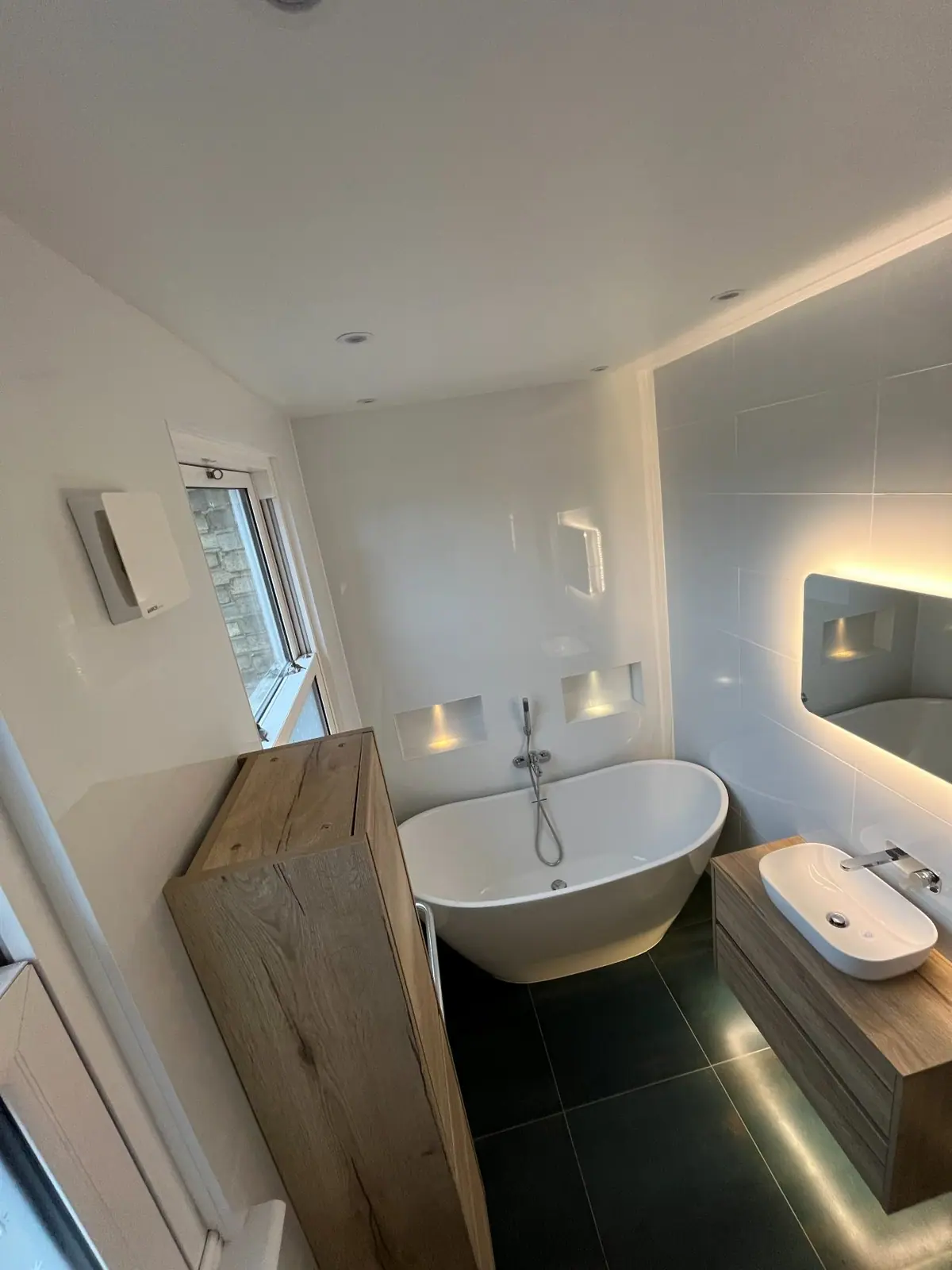 The finished bathroom with a freestanding bath, lit mirror, timber vanity unit and dark floor tiles