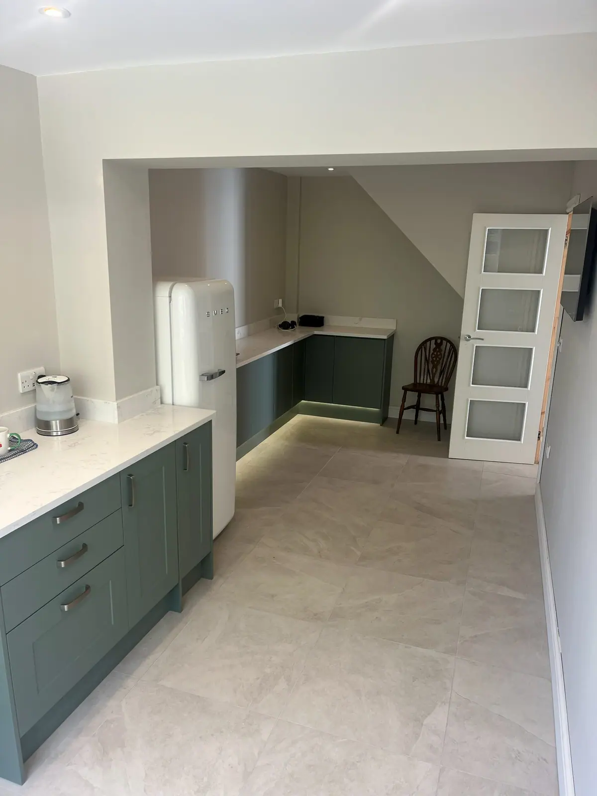 A long galley kitchen with green shaker units, a pale stone floor and plinth lighting